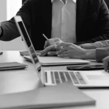 A picture of a table, laptop and notebooks, with people sitting around it and two shaking hands.